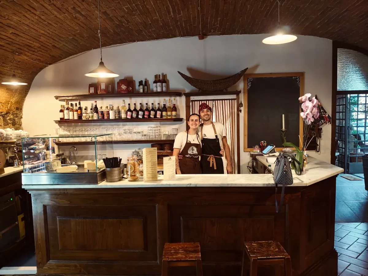 Ylenia e Niccolò dietro il bancone di legno con il piano di marmo, sotto l'arco bianco e la volta di mattoni.